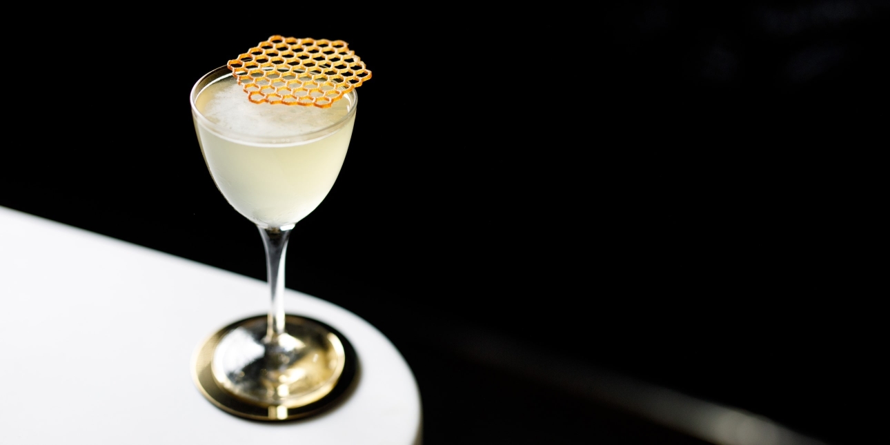 A glass of almost translucent drink with a honeycomb placed on the top, sits on a white surface. The background is black.